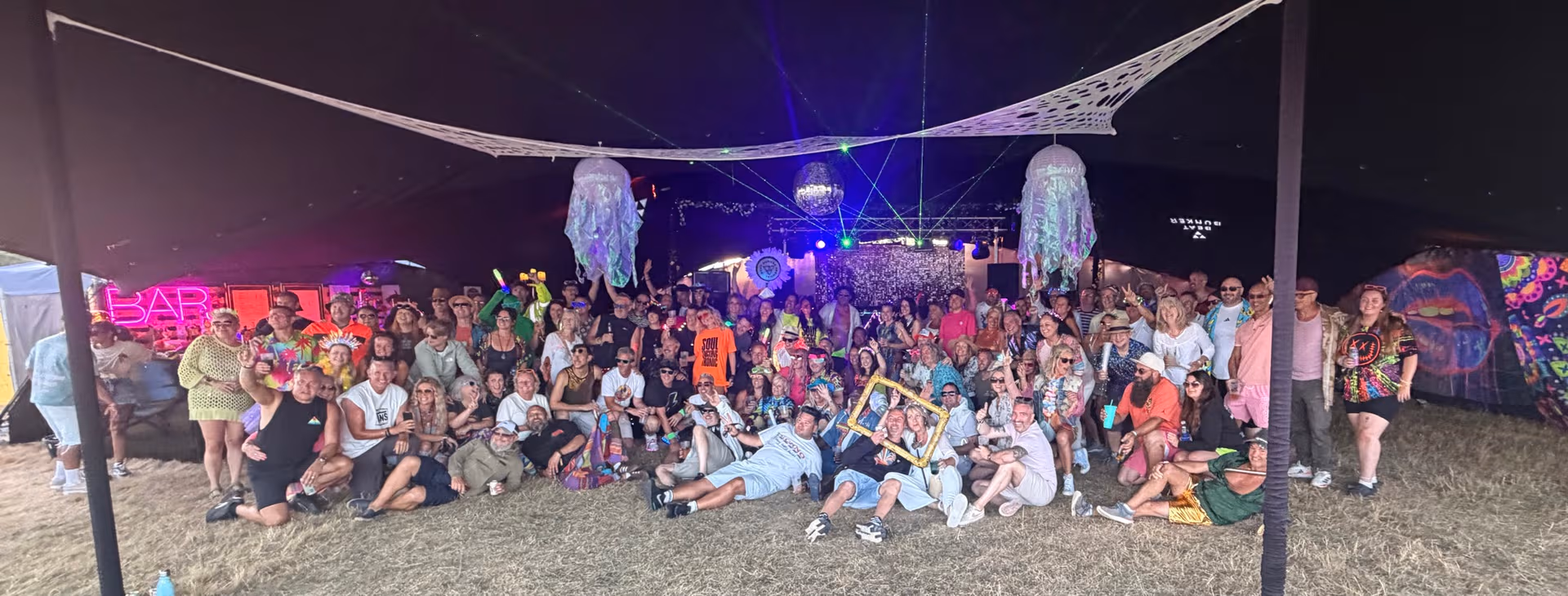 The Beat Bunker 2026 crowd posing together for a group photo under the stretch tent, lit by the bar neon, lasers and disco balls