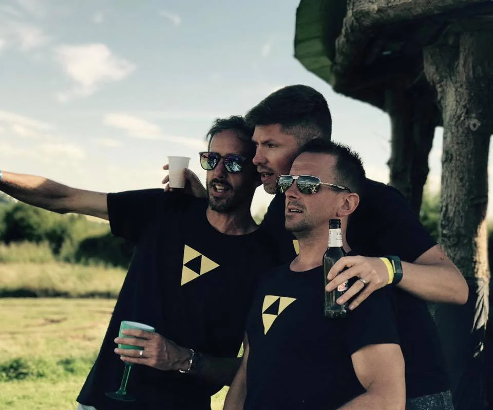 Three men wearing matching Beat Bunker shirts posing outdoors with drinks, smiling and pointing in the distance