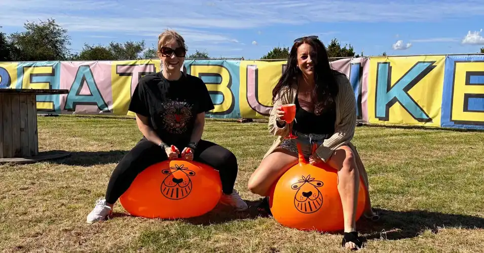 Two women sitting on orange space hoppers with drinks in hand, smiling in front of a colourful Beat Bunker banner on a sunny day